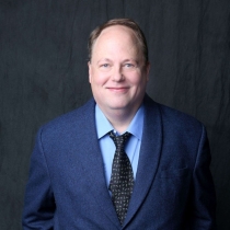 Smiling, pale, balding, heavy-set, blue-eyed, guy in a blue jacket and light blue shirt unbuttoned at the neck. His long black tie has tiny white flowers on it.