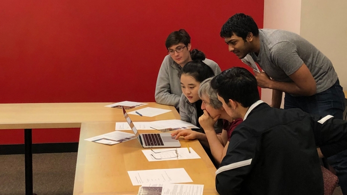 Four students look over the shoulder of an instructor at a laptop.
