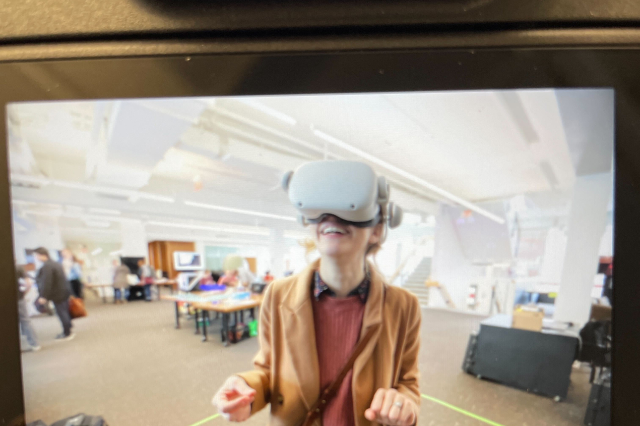 A camera takes a picture of a person wearing a red cardigan with a virtual reality headset on, looking up toward the camera in a brightly lit room