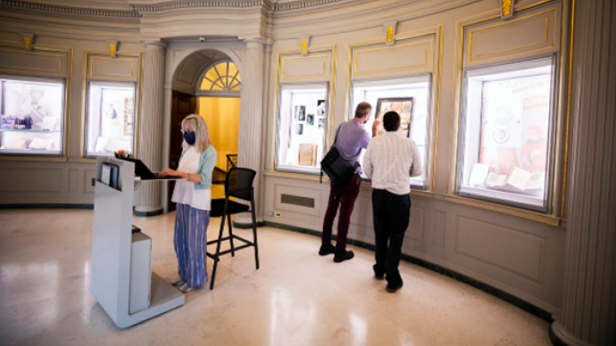 The lobby of Houghton Library, with who people looking away from the camera at a display, and one person standing and looking toward the camera at the front desk