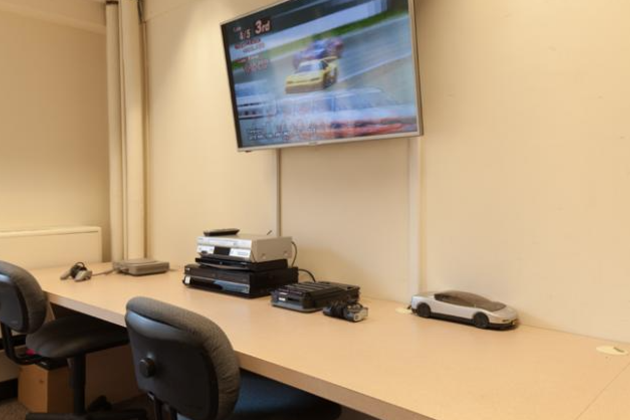A room is shown with a long desk with multiple media consoles, and a tv mounted above the desk on the wall