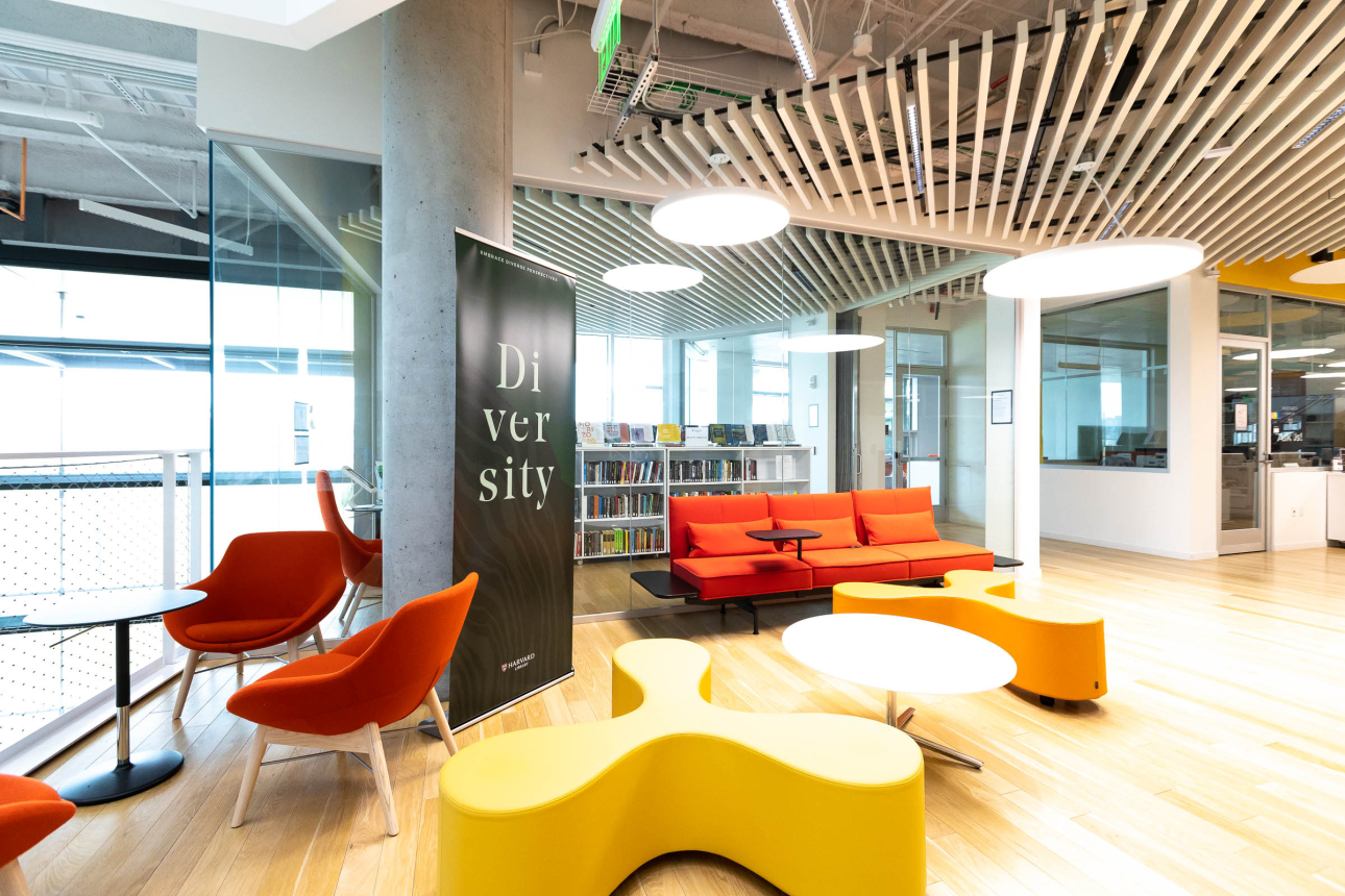 Modern library lounge with bright orange and yellow seating, round white tables, a bookshelf wall, and a large “Diversity” banner inside Harvard’s Science & Engineering Complex Library.