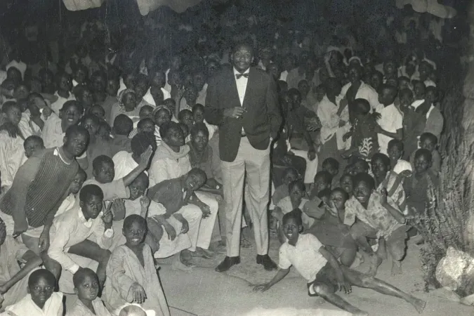 A tall man wearing a suit stands in a room filled with smiling children.