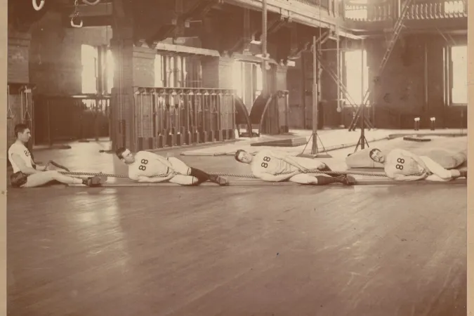 Black and white photo of male students on a gymnasium floor pulling on a rope