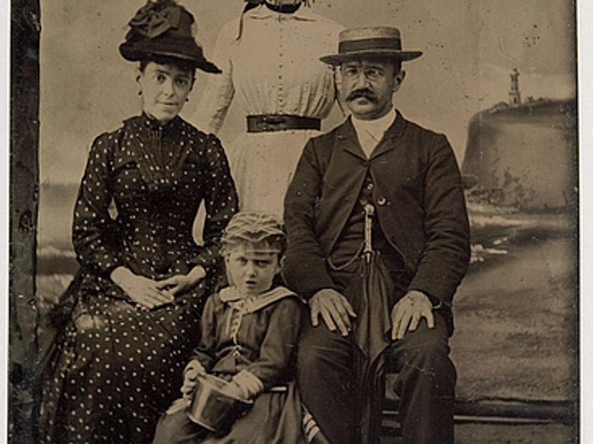 Sepia toned tintype photo of two women, a small girl, and a man. A face of the standing woman is scratched.