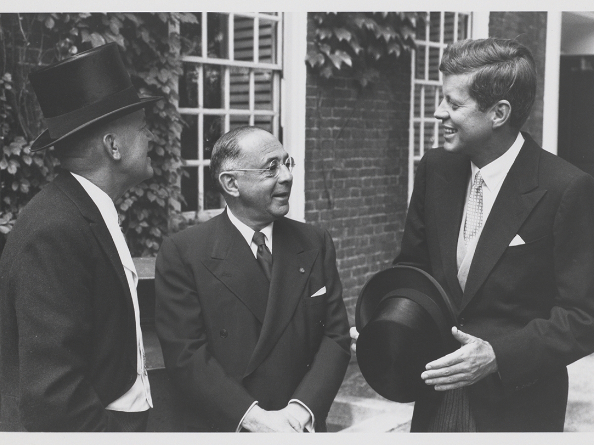 Three men standing in front of a brick building. John F. Kennedy stands at the right, smiling, holding a hat.