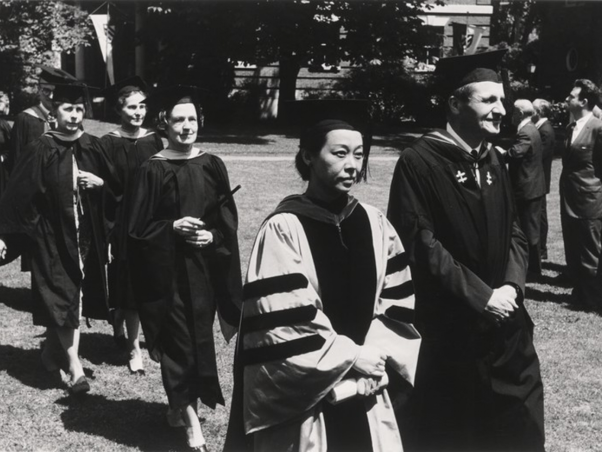 An outdoor commencement procession of faculty and administrators. Rulan Chao Pian, wearing Harvard's crimson doctoral regalia, is walking next to a male faculty member in black regalia, followed by several women in generic black robes.