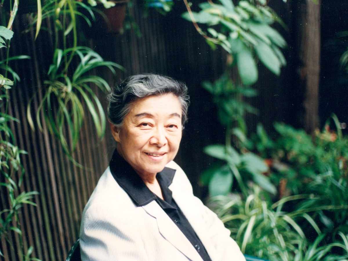 Rulan Chao Pian, sitting outside surrounded by lush greenery, and smiling at the photographer. She is wearing a black collared shirt and cream blazer, and her greying dark hair is pulled back.