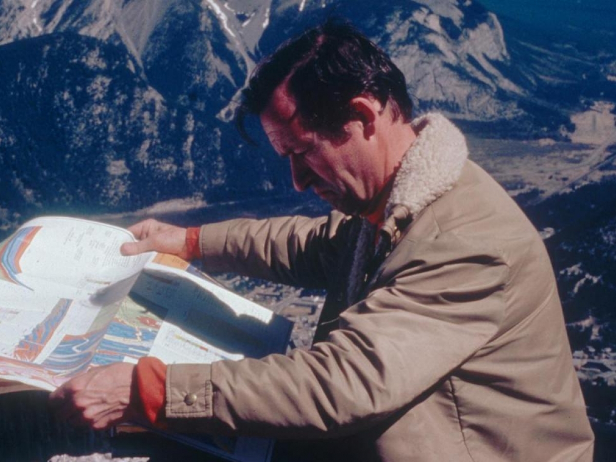 A middle-aged man in a tan coat stands outside, studying a map. Snow-capped mountains can be seen in the distance.