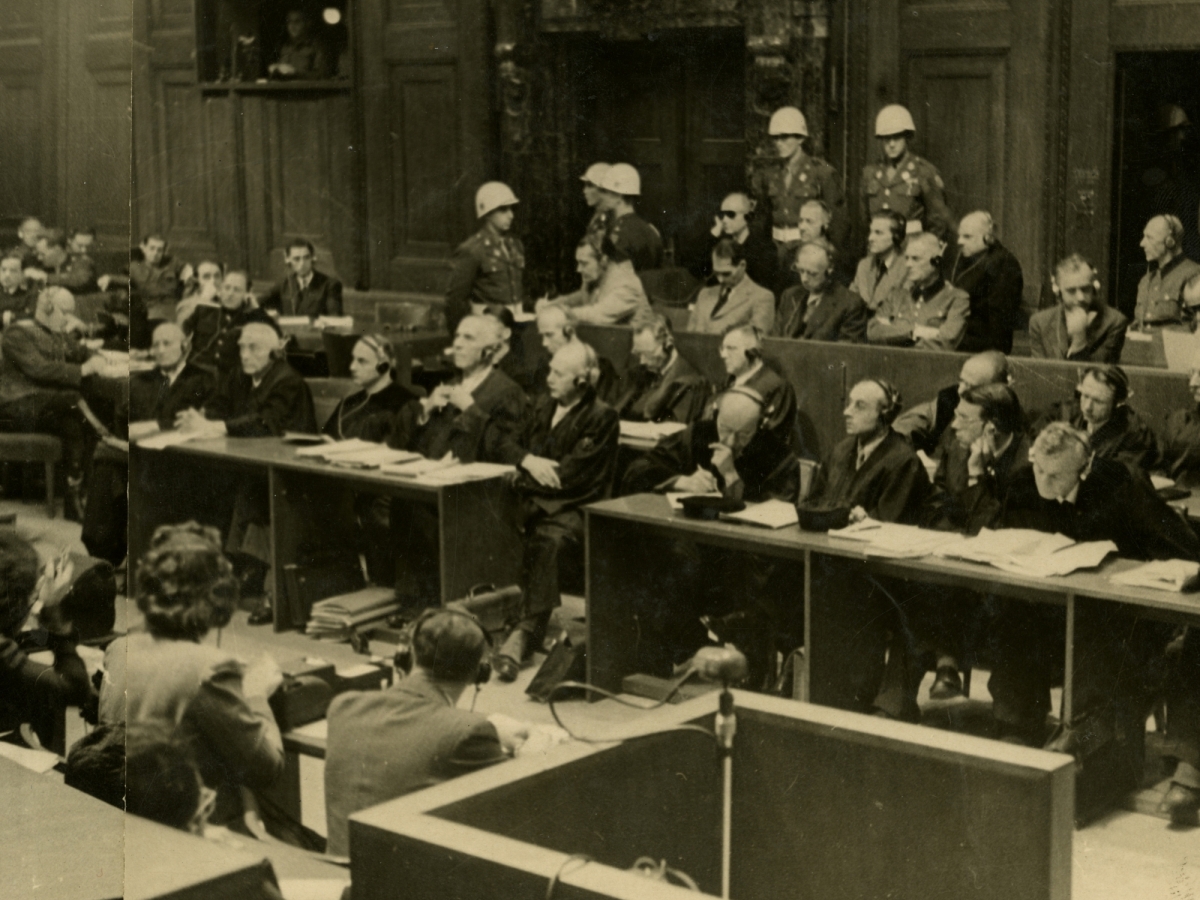 Black and white photograph showing the prisoner’s dock in the Nuremberg trials courtroom, with the accused seated in a row with military guards wearing white helmets standing behind them. 