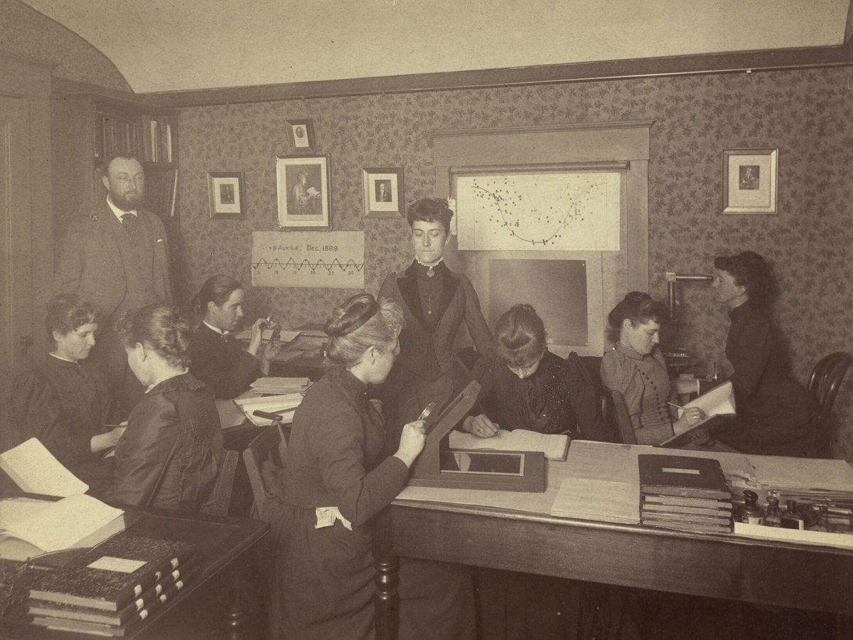 Photo of eight women and one man in a classroom. Maury is seated in profile, third from left, peering into a magnifying glass.