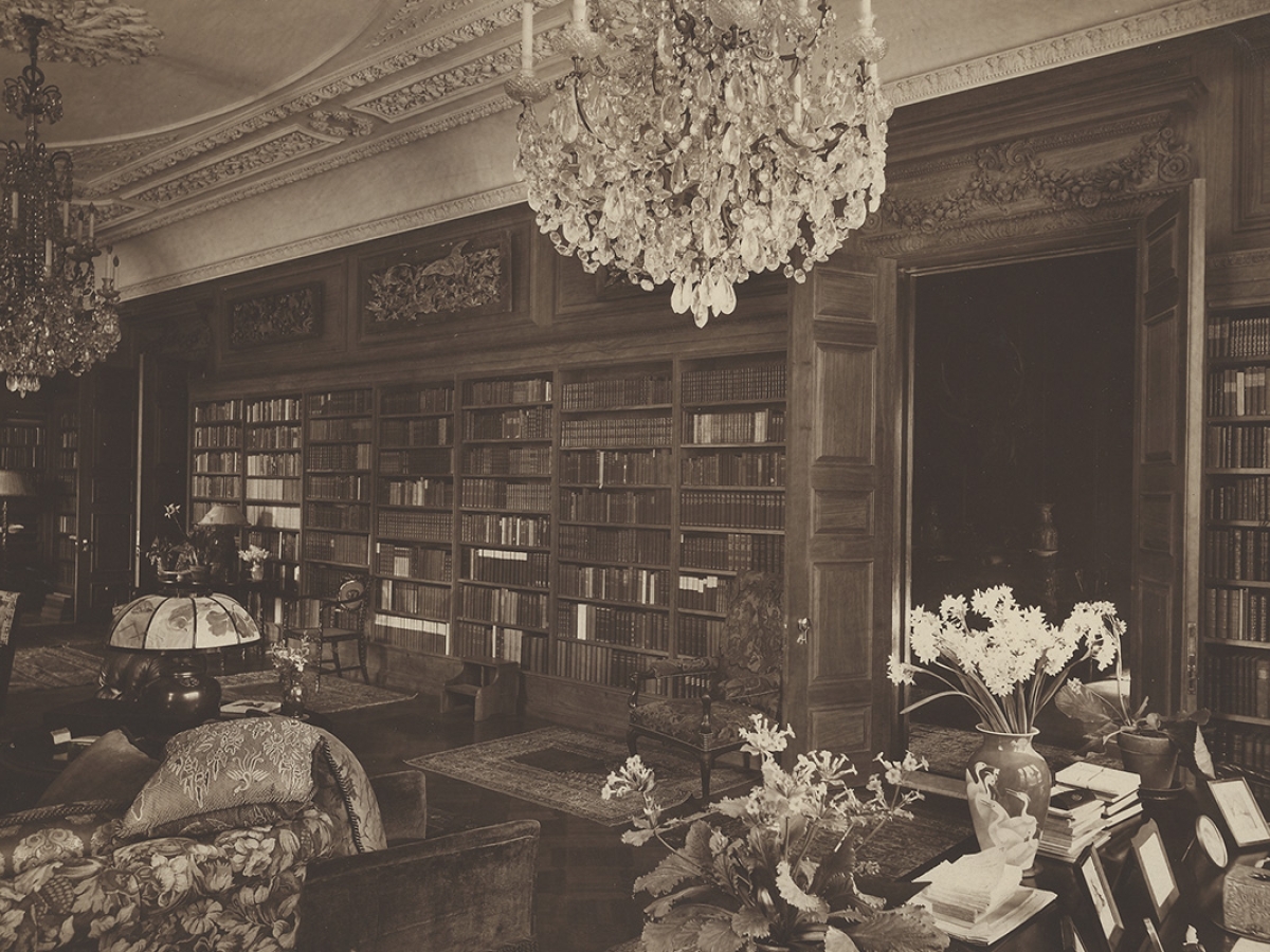 A sepia off-center photograph of Lowell’s library at Sevenels with her desk on the right and two chandeliers hanging in frame.