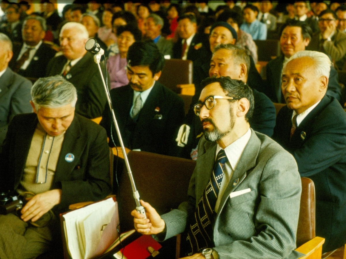 Siberian-born ethnomusicologist Eduard Alekseyev, wearing a gray suit and a spotted tie, holds an audio recording device.
