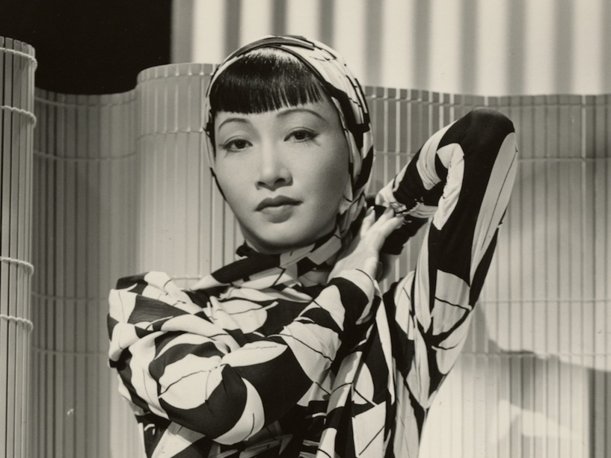 woman in dress with stylized leaves and head scarf poses in front of bamboo screen.