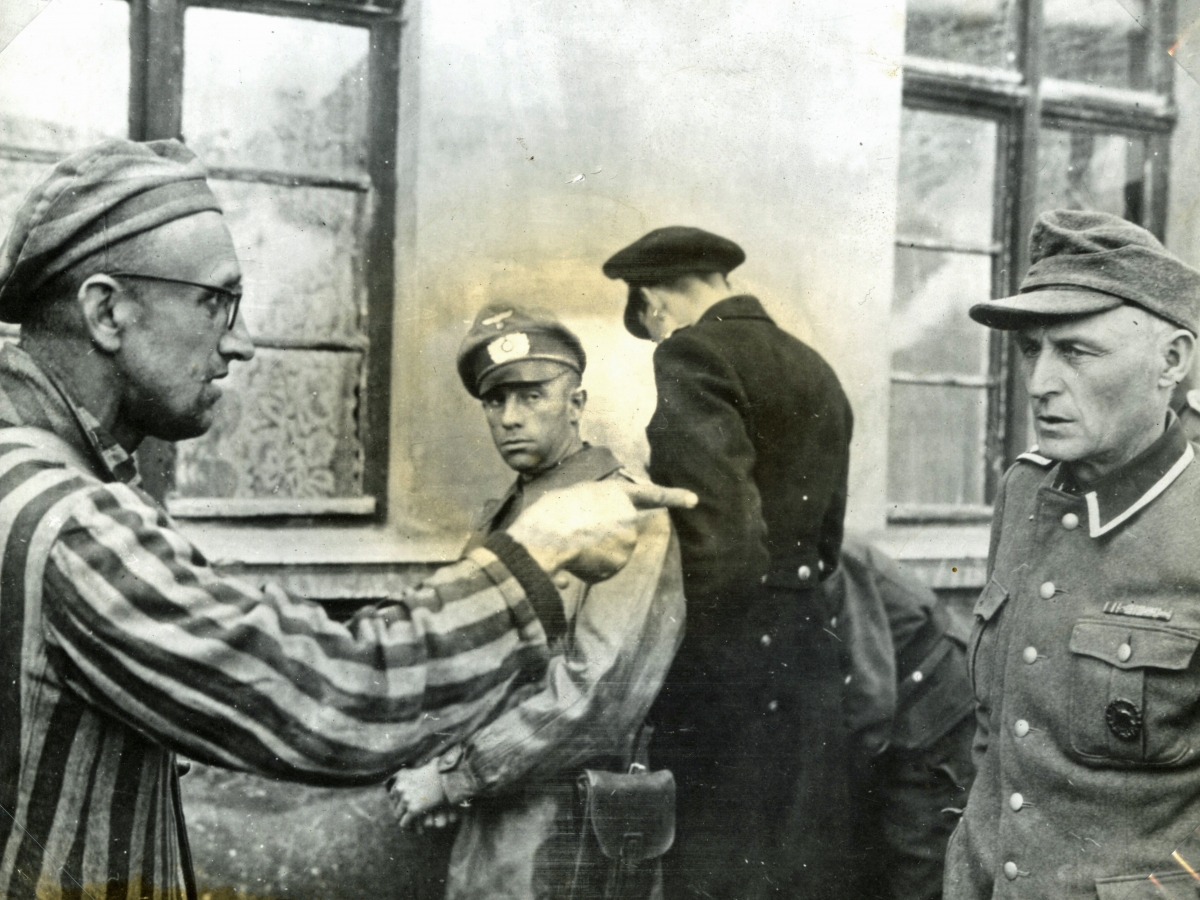 Black and white photograph showing a concentration camp prisoner at Nordhausen-Dora wearing a striped uniform and cap, pointing a finger accusingly at a SS guard, with two guards in the background. 