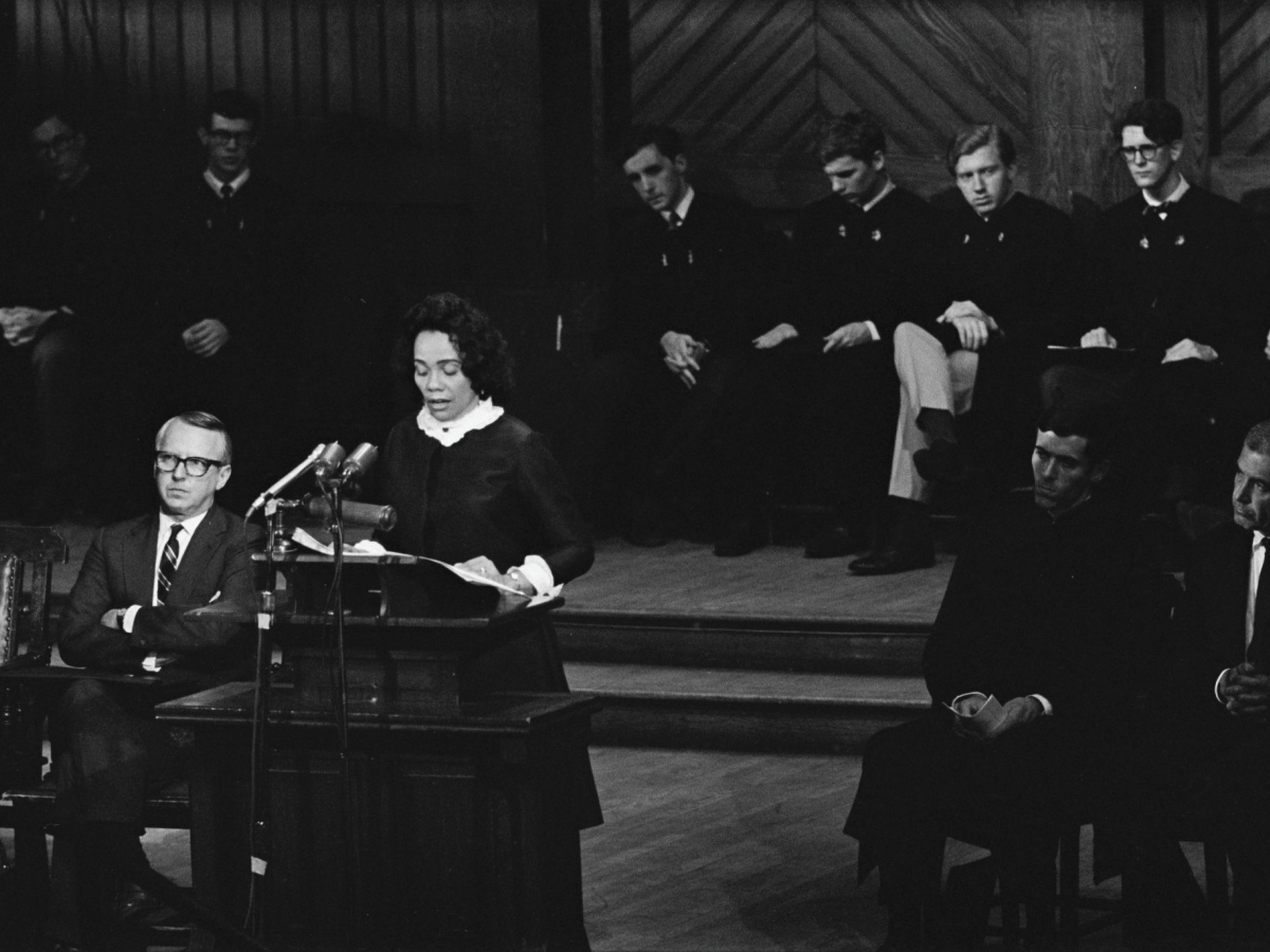 Woman speaking at a lectern with men sitting on a dais all around her.
