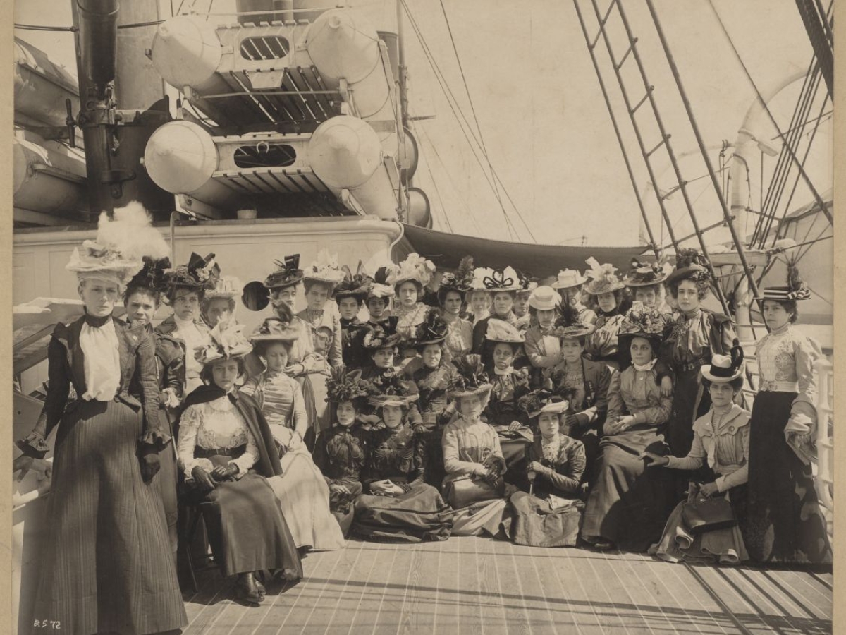 Women in formal dress and hats on the deck of a U.S. naval ship.