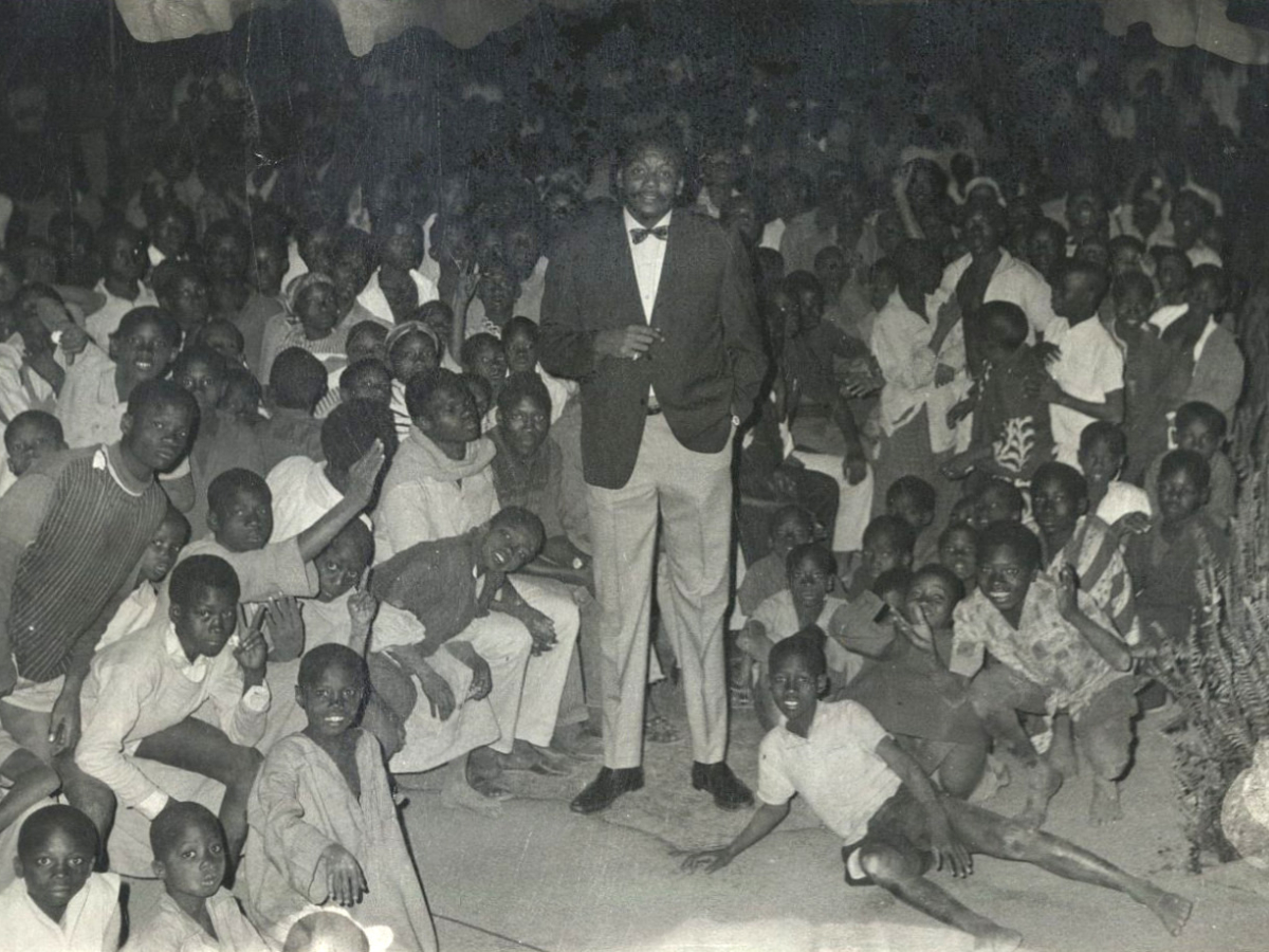 A tall man wearing a suit stands in a room filled with smiling children.