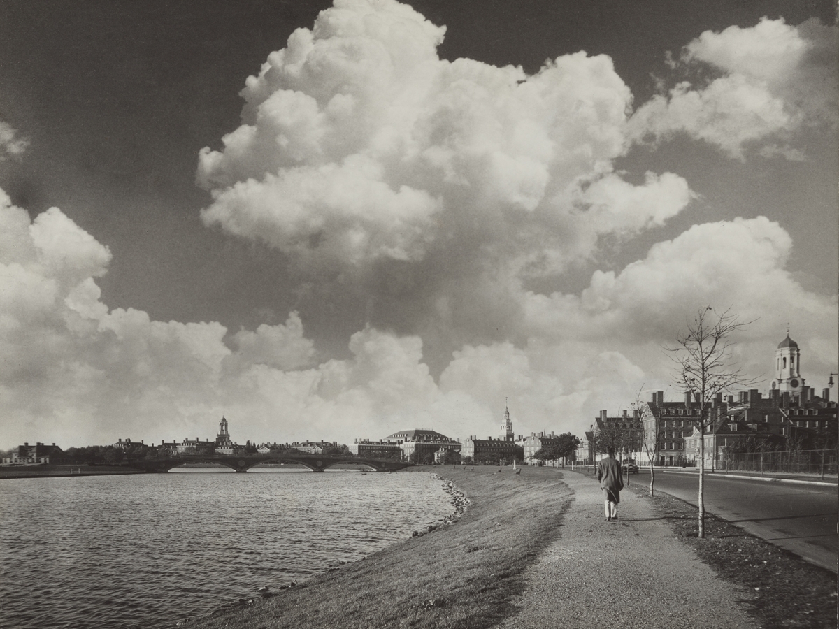 A person walks down the sidewalk alongside Memorial Drive. Several towers on the Harvard upperclass houses appear in the background. The Charles River runs along the left.