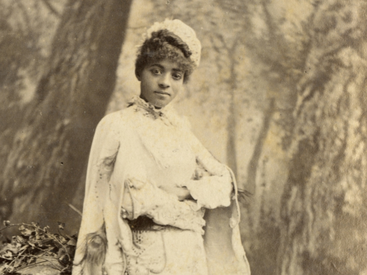 Black woman in a white dress with cape, white bonnet, and gloves stands before a backdrop painted with a forest scene.