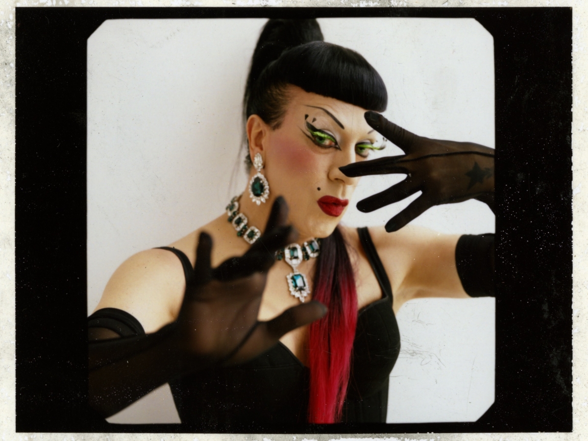 A drag performer with long black and red hair, over-the-elbow black gloves, black dress, and chunky jewelry with her hands posed in front of her face.