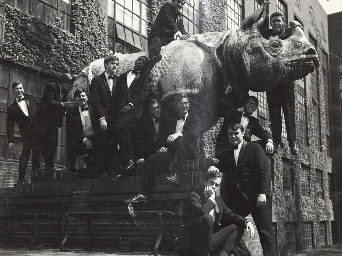 Young men in tuxedos stand on statue of rhino outside of ivy-covered building