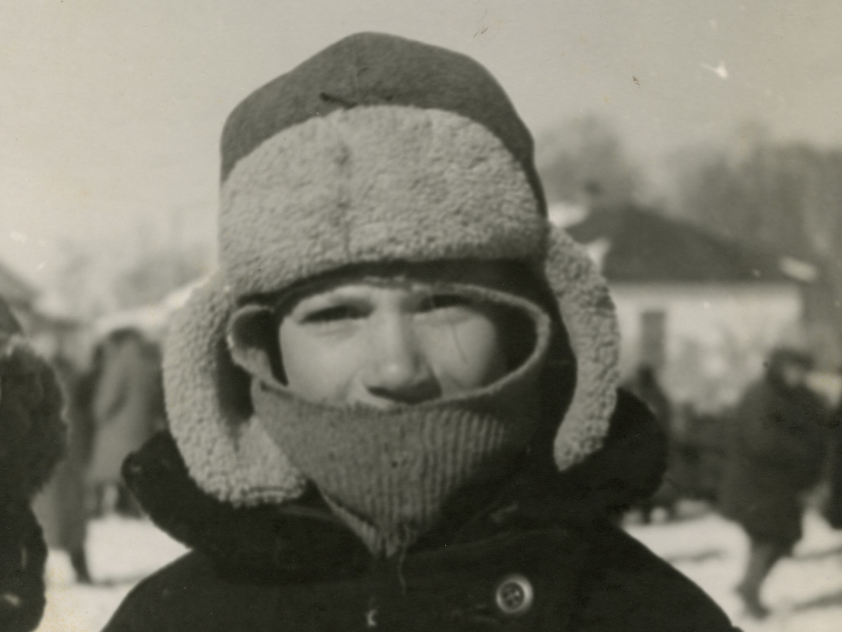 A small boy wearing a cap with earflaps and a muffler outside on a bright snowy day with a crowd of people around him.