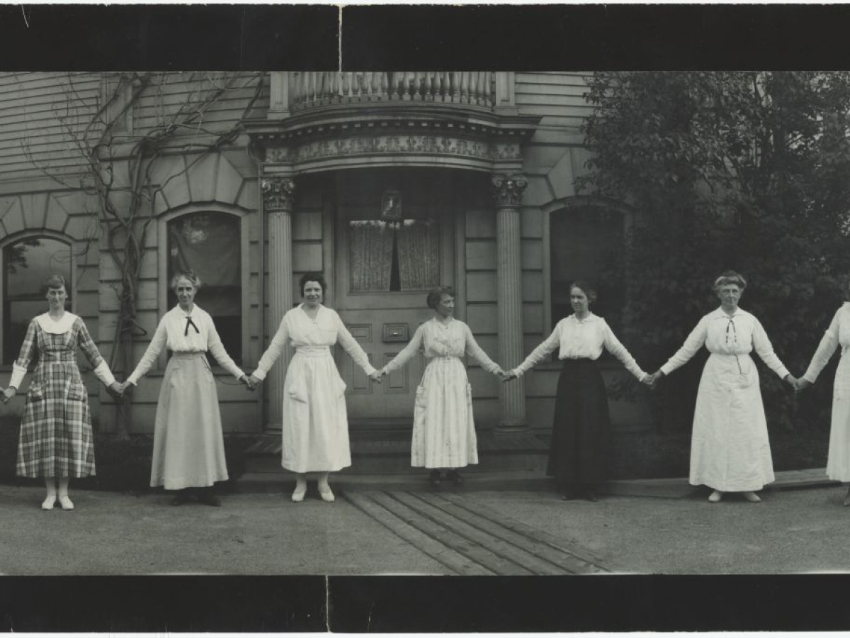 Women and men stand in front of a building in a "paper doll pose," holding hands with arms stretched beside them.