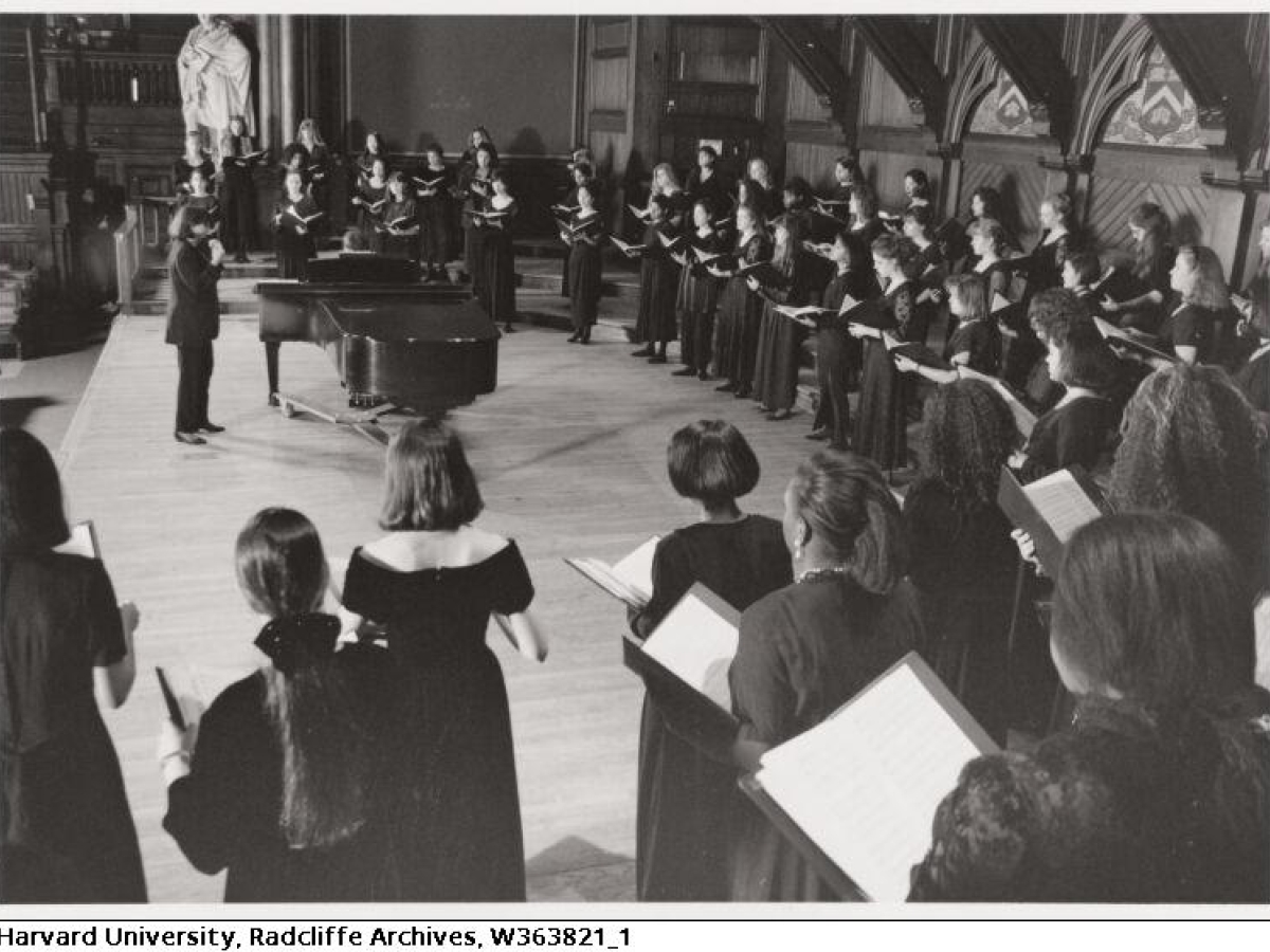 A chorus of several dozen people in floor length black dresses, standing in an arc around a conductor by a grand piano.