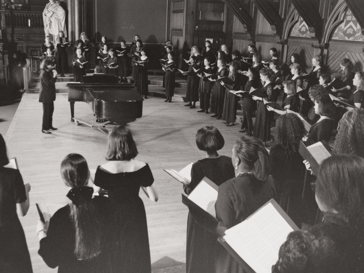A chorus of several dozen people in floor-length black dresses, standing in an arc around a conductor by a grand piano.