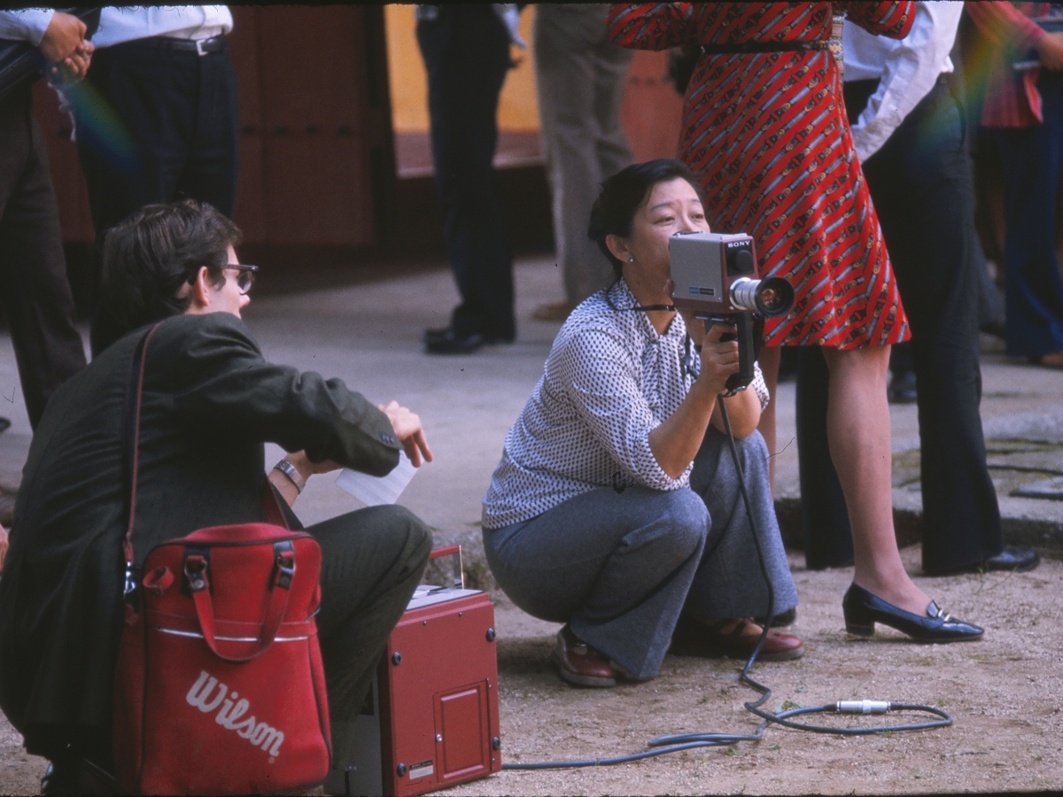 A crouching woman with an attentive expression, holding a video camera, amidst a group of standing observers.
