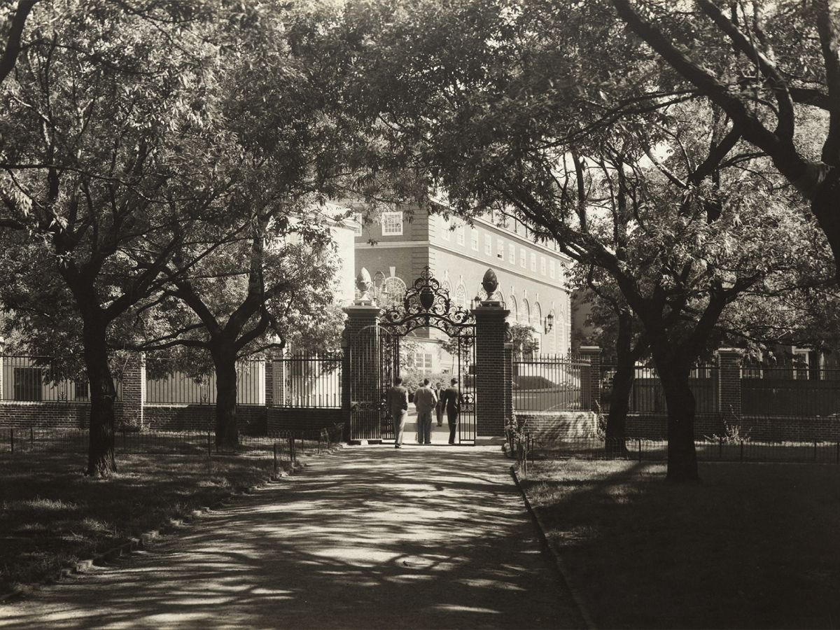  Three young men exit through a gate, toward several buildings. Elm trees throw dark shadows on the grounds around them.