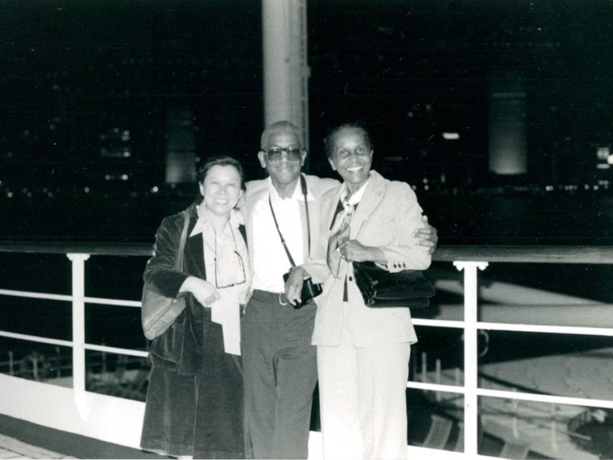 Three people standing close to one another and smiling widely at the photographer. The man in the center of the group has a camera around his neck, and the two women are carrying shoulder bags. They are outside at night, in front of a metal railing.
