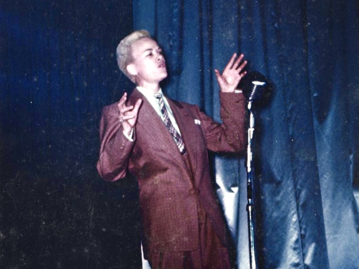 Male impersonator in a brown suit and tie sings into microphone in front of a blue stage curtain.
