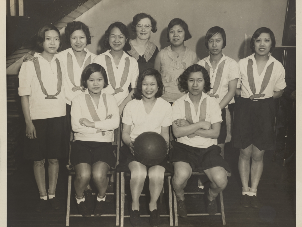 A black and white photograph of the members of a Chinese American girls basketball team, gathered togehter for a team photograph.  The three girls in the front row are seated with the center girl holding a basketball. The other five girls are standing behind them. The girls are in their basketball unifroms, dark skirts and white blouses with a ribbon draped aroud their shoulders. Two teachers or coaches are in the back row center.  