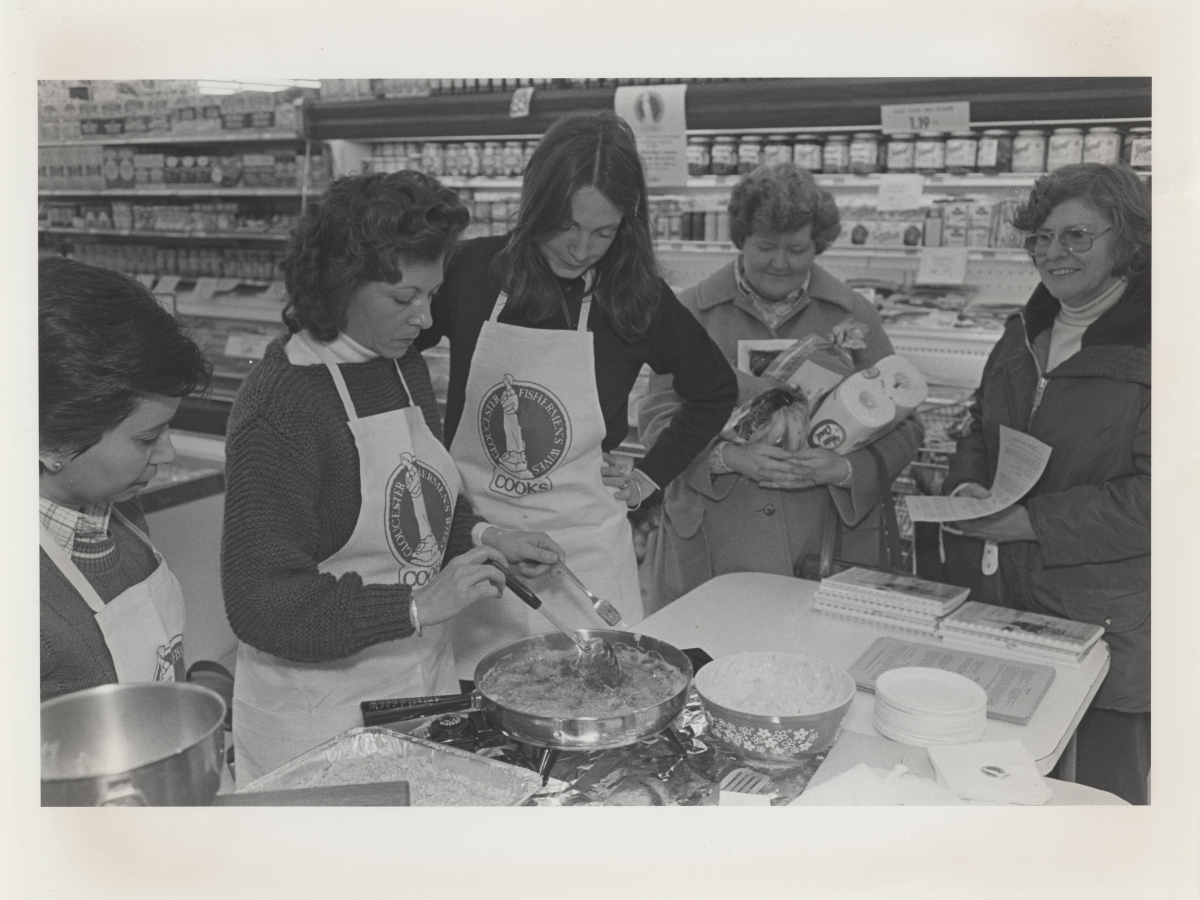 Women from the Fishermen's Wives of Gloucester giving a cooking demonstration at a grocery store. Three women in white aprons standing around a white folding table. One of the women is cooking fish in a frying pan. Two women customers are in the background watching. There are also stocked shelves in the background.