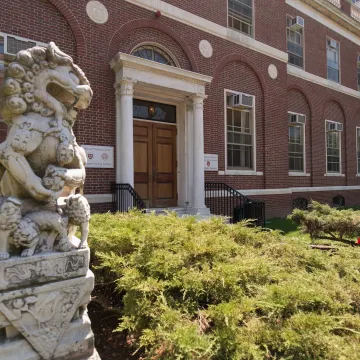 Exterior entrance of the Harvard-Yenching Library, a red brick building with a columned doorway, flanked by stone guardian lion statues and landscaped greenery in the foreground.