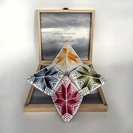an open container with a chatterbox game inside. Chatterbox, a folded paper structure has embroidered tatreez (traditional Palestinian cross-stitch) on the outside.