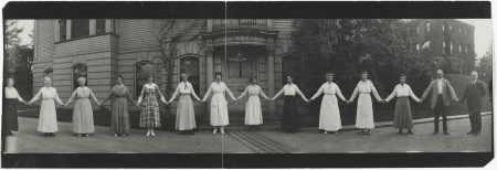 The staff of the Harvard College Observatory stand in a line holding hands in a panoramic snapshot.