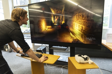 A man plays a video game, and on the large screen TV in front of him we can see the tunnels of an an Egyptian tomb