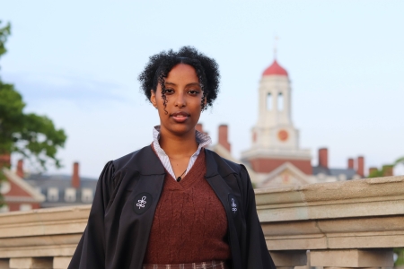 Birukti Tsige, wearing a red sweater under a black graduation robe, stands on a bridge over the Charles River