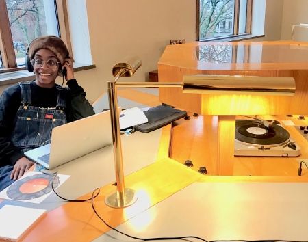 An individual sits at a computer in the Woodberry Poetry Room, smiling at the camera, a record player can be seen in the righthand side of the photo