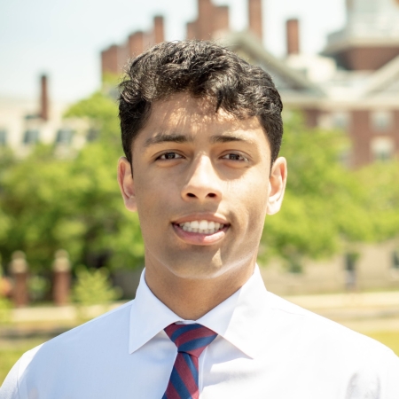 Ishaan Prasad, standing outdoors, and wearing a white collared shirt and striped tie, smiles at the camera