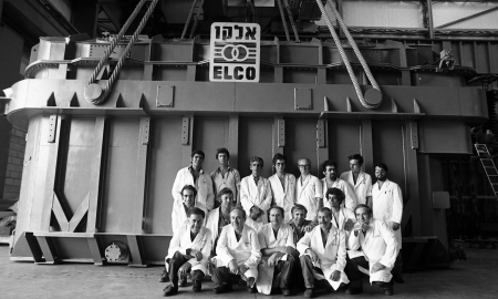 Seventeen men in white lab coats stand in front of a large metal electricity generator with a sign reading "Elco" above their heads.