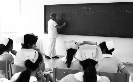 A teacher wearing white stands at a blackboard in front of a class of women wearing nurse's caps. The words on the blackboard relate to pregnancy and conception.