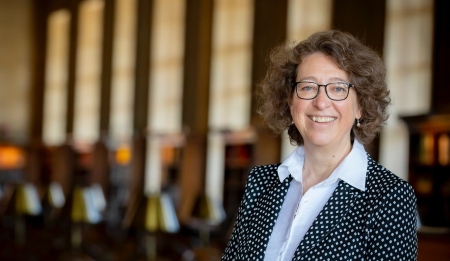 Martha Whitehead stands in front of windows, smiling and wearing a collared shirt and glasses