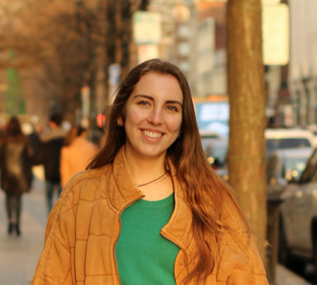 Tess Kelley, a white woman with brown hair, smiles facing the camera. She is wearing an orange coat and a grey top underneath