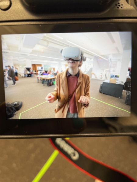 A camera takes a picture of a person wearing a red cardigan with a virtual reality headset on, looking up toward the camera in a brightly lit room