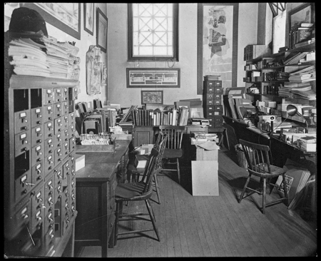 A black and white photo of an old room with a card catalog and desks stacked with books
