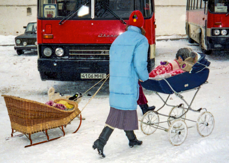 A woman pushes a stroller and pulls a sled by buses parked on a snowy street.